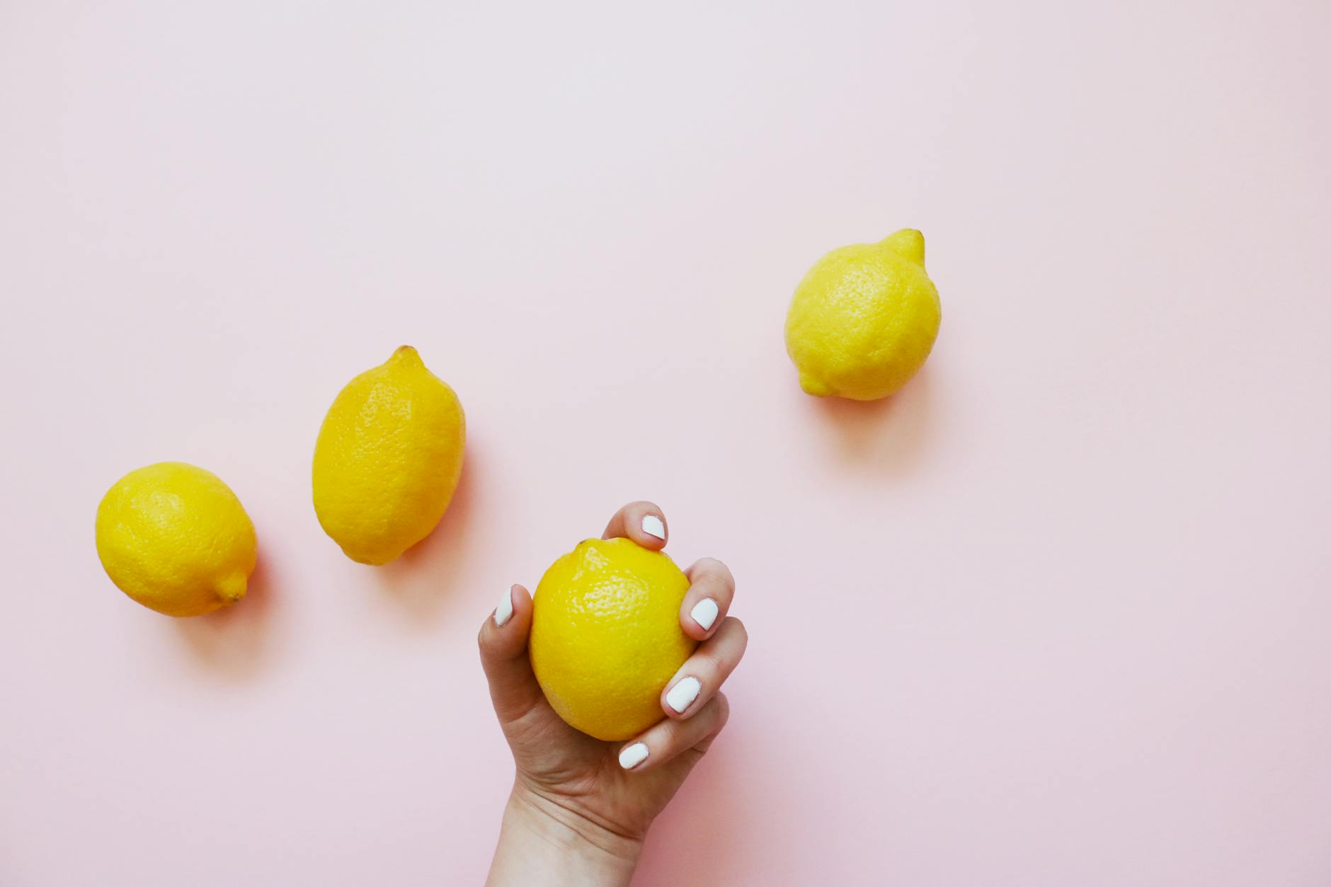 A hand holding a fresh yellow lemon on a soft pink background, surrounded by additional lemons.