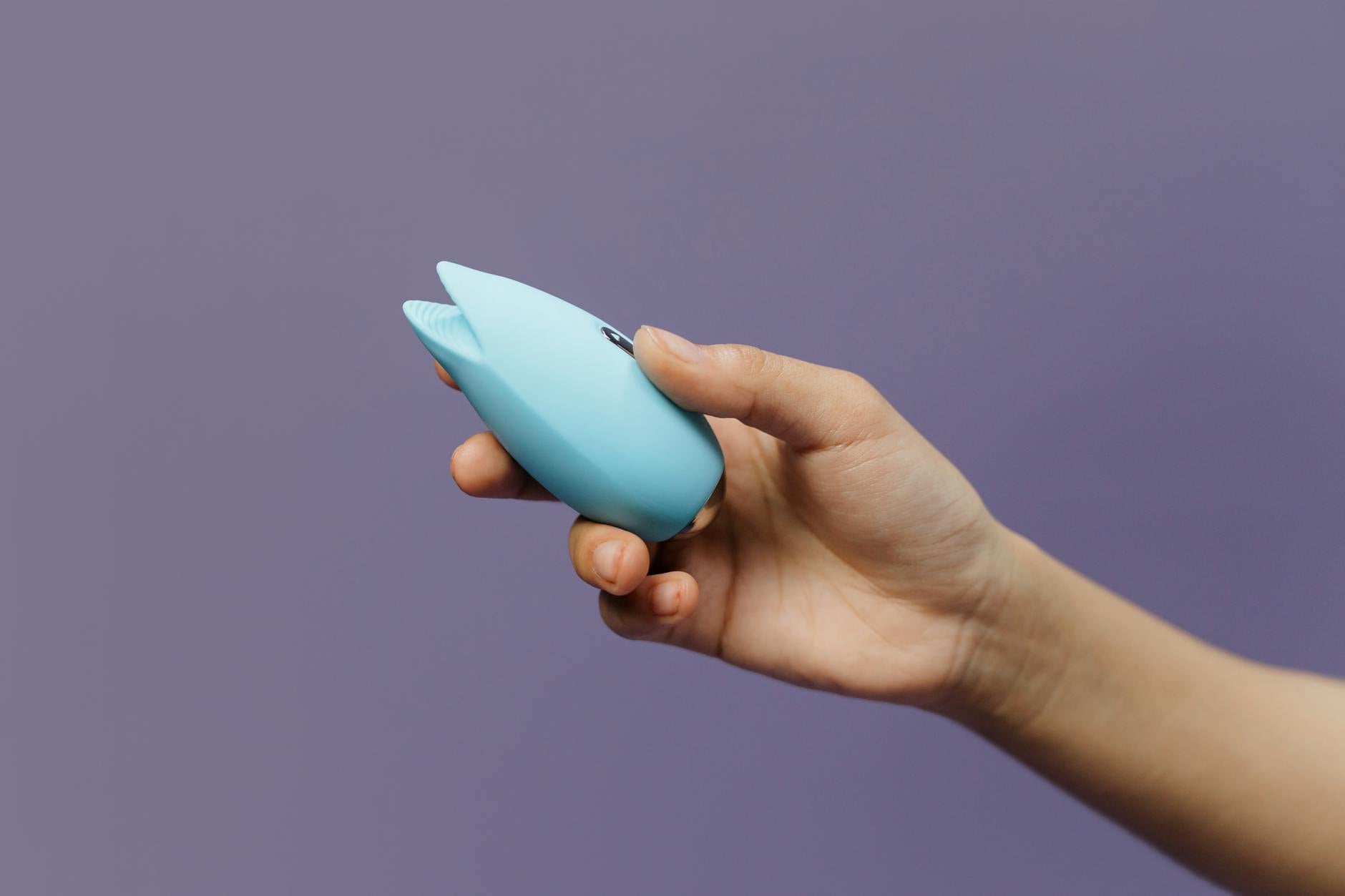 A hand holding a blue clitoral vibrator against a purple background, symbolizing sexual wellness and self-care